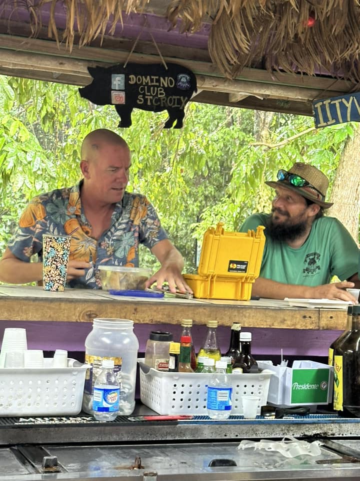 Two men chatting at the Domino Club bar with a pig-shaped sign overhead and bottles lining the counter