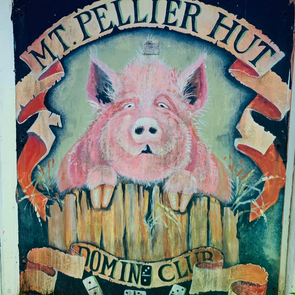 The iconic hand-painted Mt. Pellier Hut Domino Club sign featuring a pig leaning over a wooden fence, with domino tiles scattered below
